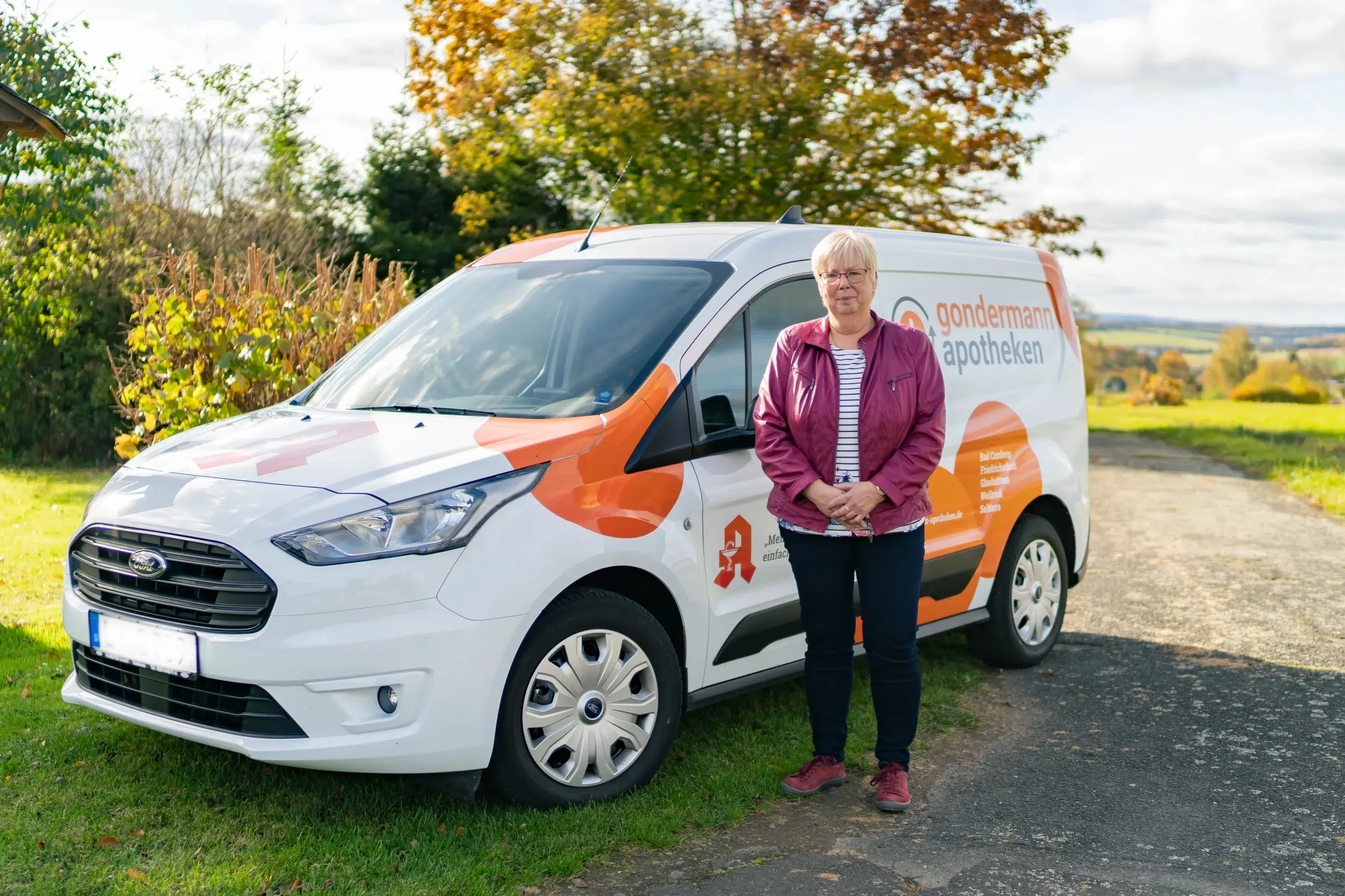Symbolbild für den Apotheken-Botendienst: Eine Botendienstfahrerin posiert im Grünen vor dem Fahrzeug – zuverlässige Medikamentenzustellung mit persönlichem Service direkt nach Hause